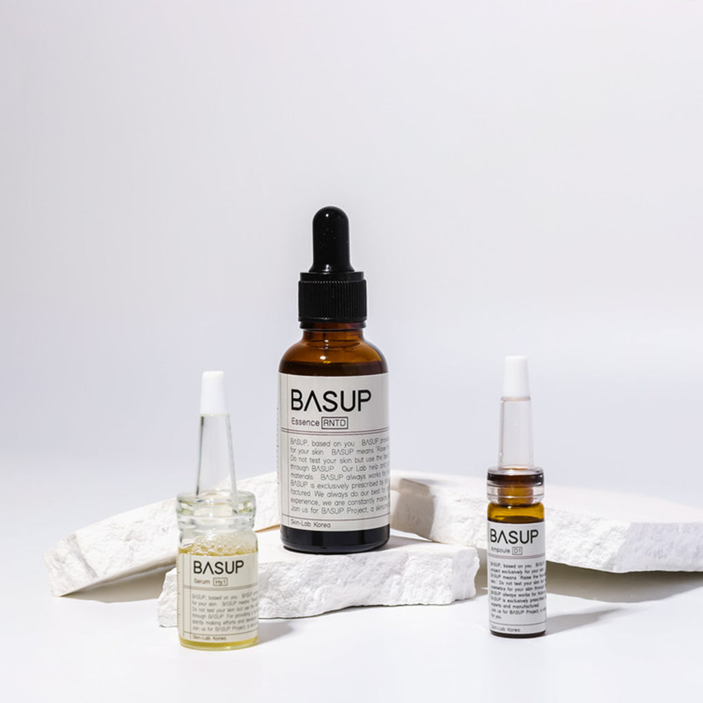 BASUP essence in an amber dropper bottle with two small ampoules displayed on white stone slabs.